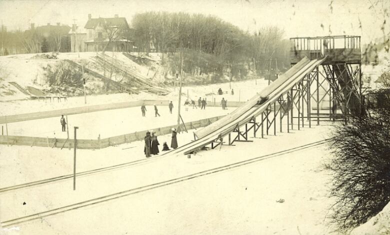 Black and white photo of a tall, wooden toboggan slide on a frozen river, next to a hockey rink.