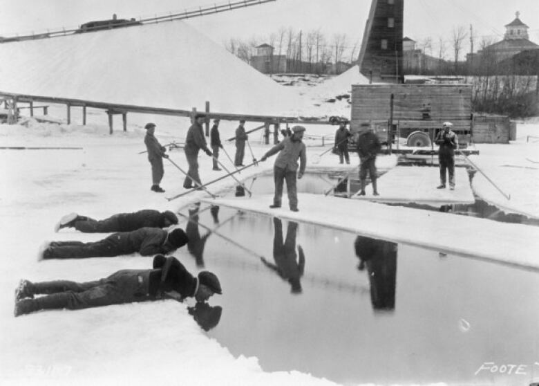 Black and white photo shows three people lying on their stomachs on ice, and leaning into an open space of water to drink from it.