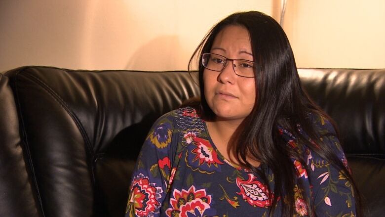 A woman with long black hair in a floral shirt sits on a black couch.