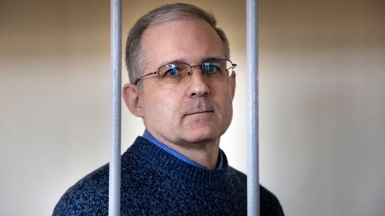 A picture of a man, with grey hair and wearing glasses, standing behind two metal prison bars.