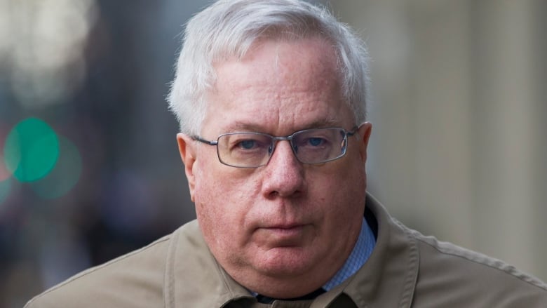 A man in a trench coat and glasses squints at the camera. 