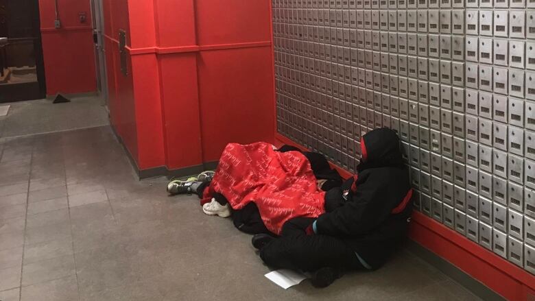 Two people, with their backs to the camera, huddle on the floor next to a wall of post office boxes.