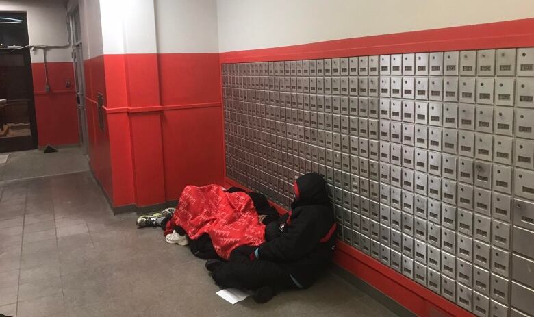 Two people, with their backs to the camera, huddle on the floor next to a wall of post office boxes.