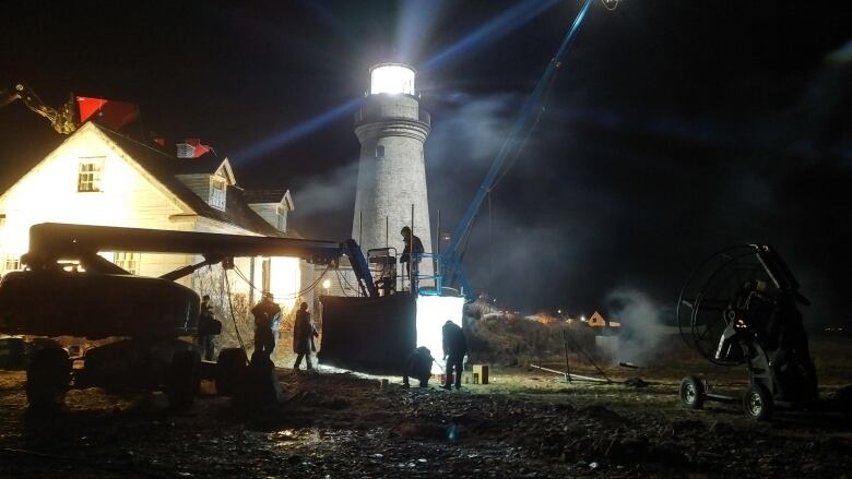 A film crew work in front of a lighthouse on a dark night.