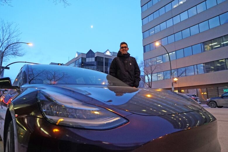 A man stands in downtown Saskatoon beside a Tesla car.