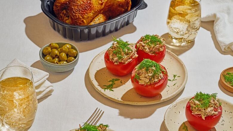 3 chicken salad stuffed tomatoes on a beige plate. They're sitting on a light beige tablecloth with more plates, a small bowl of green olives, and a rotisserie chicken arranged around them. 