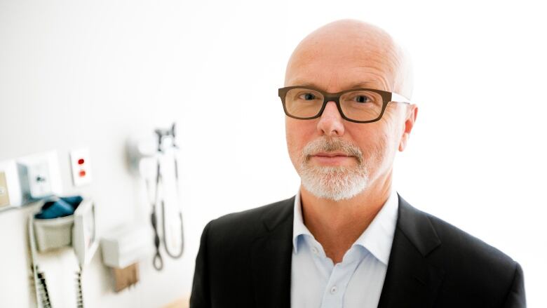 Bearded man with glasses in a business suit, standing in a medical office. 