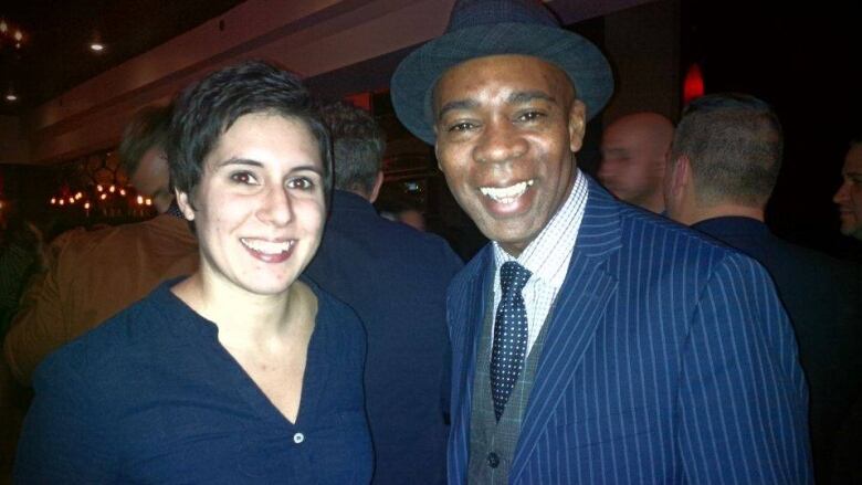 A woman in a blue shirt and a man wearing a suit and fedora smile for the camera