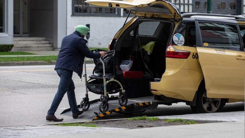 A man wearing a turban wheels a wheelchair into the back of a yellow taxicab.