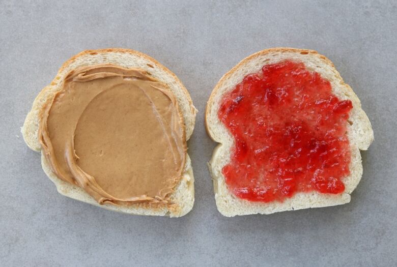 Two pieces of white bread lay side by side, one spread with peanut butter, the other with jelly.