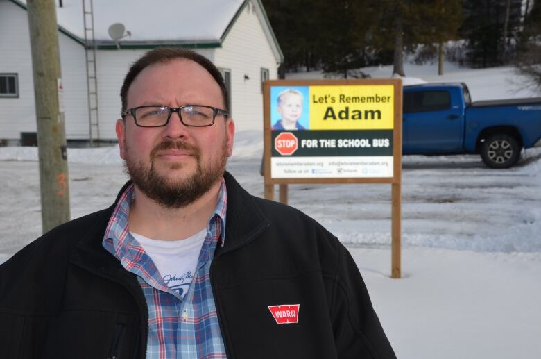 A man with a beard standing in front of a sign that says Let's Remember Adam.