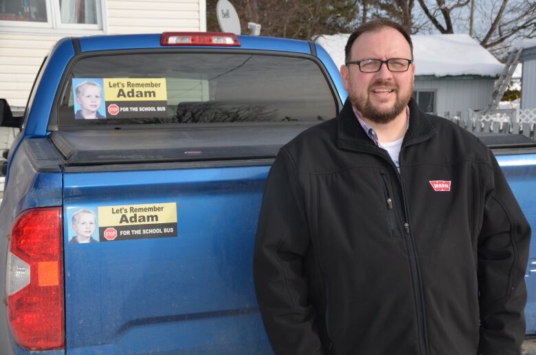 A man stands in front of a blue pickup truck with a Let's Remember Adam bumper sticker.