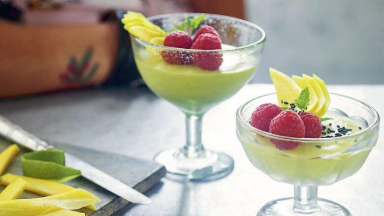 2 stemmed glass dishes with a yellow pudding in them and raspberries on top, sitting on a table. a person's arm, who is sitting at the table, is visible in the background. 