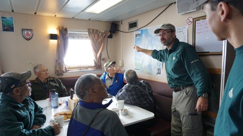 in a room on a ship, a man stands at the front of an points to a map while a group of Inuit elders sit around a table and listen