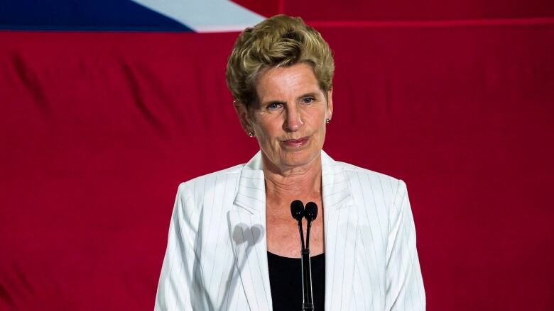 A woman with short hair in a white blazer stands at a microphone.