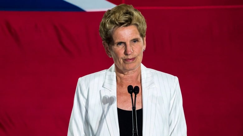 A woman with short hair in a white blazer stands at a microphone.