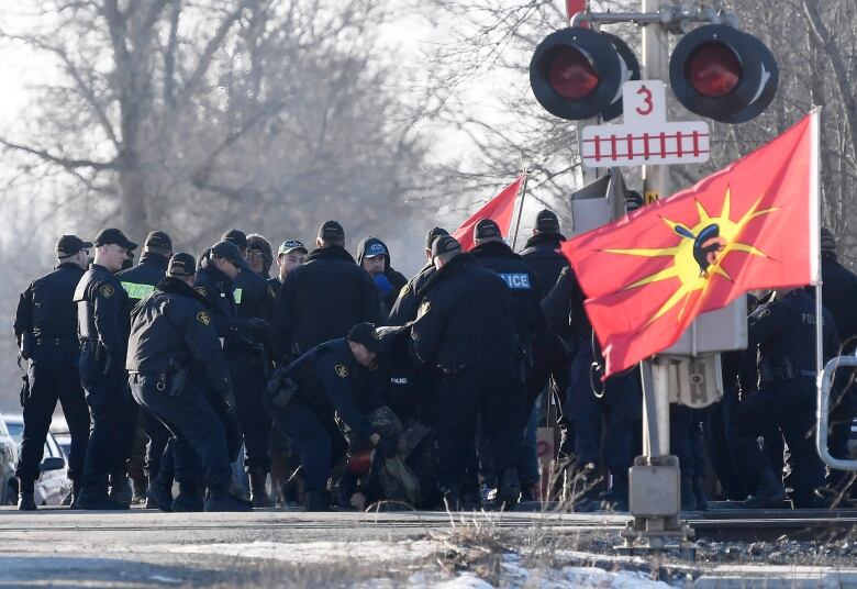 Police surround someone next to railroad tracks in winter. There's a red flag with a warrior logo nearby.