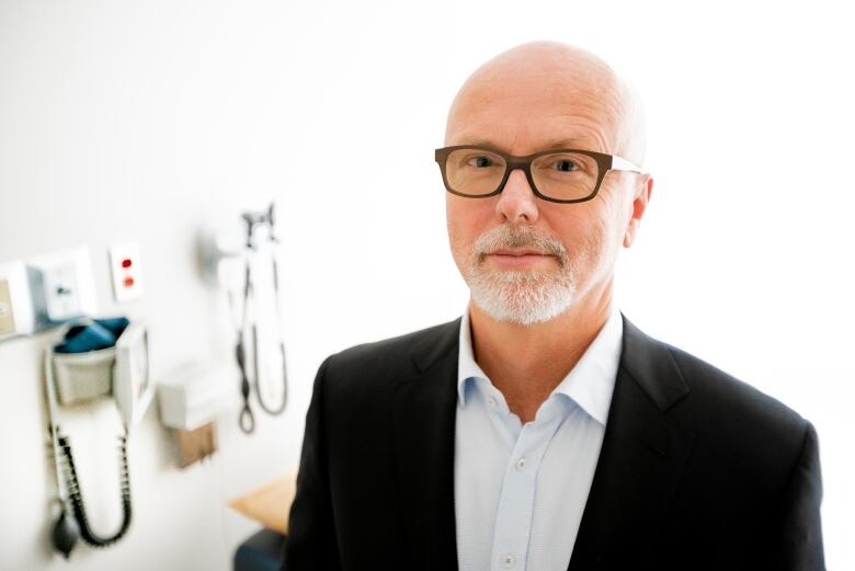 A bearded man with glasses stands in a doctors office looking at the camera.