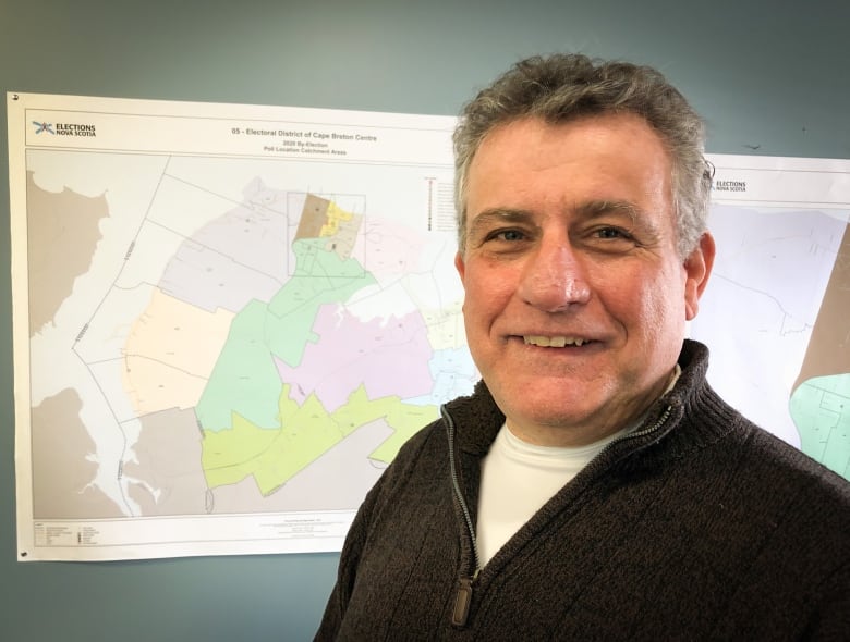 A man with a dark sweater and white t-shirt smiles with a map behind him.