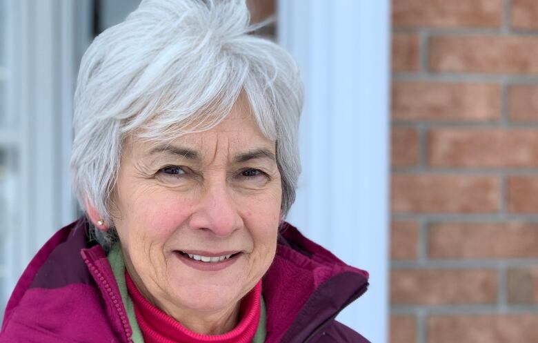 A woman with short white hair and a pink jacket smiles at the camera.