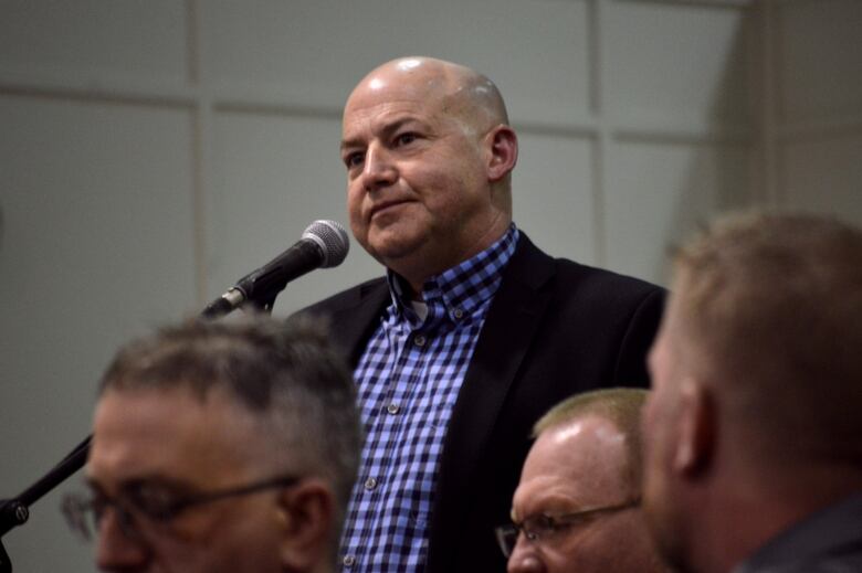 A white, bald man in a checkered shirt and a dark-coloured jacket. He's standing at a microphone.