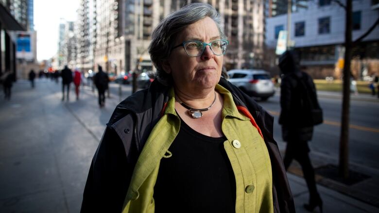 A woman with grey hair, blue glasses, a green shirt and a black jacket stands on the sidewalk of an urban street