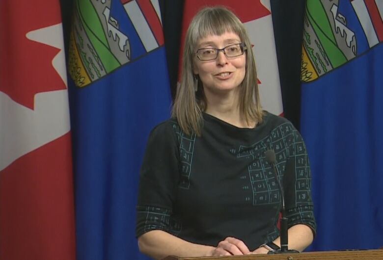 A woman in long hair and glasses stands in front of Canadian and Albertan flags.