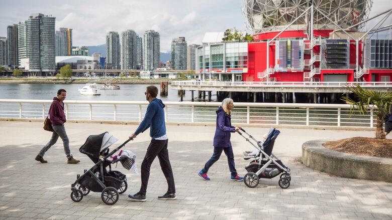 Two people push strollers with Science World in the background while another person walks by. 