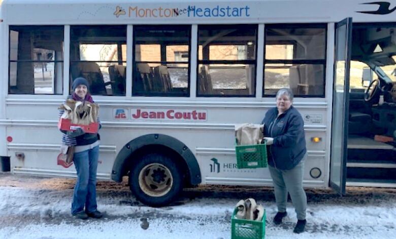 Two women stand in front of a bus handing out school lunches.