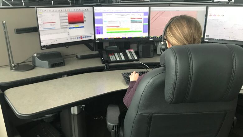 A woman is seated in a chair with headphones on, while looking at four computer monitors to help her respond to emergencies. 