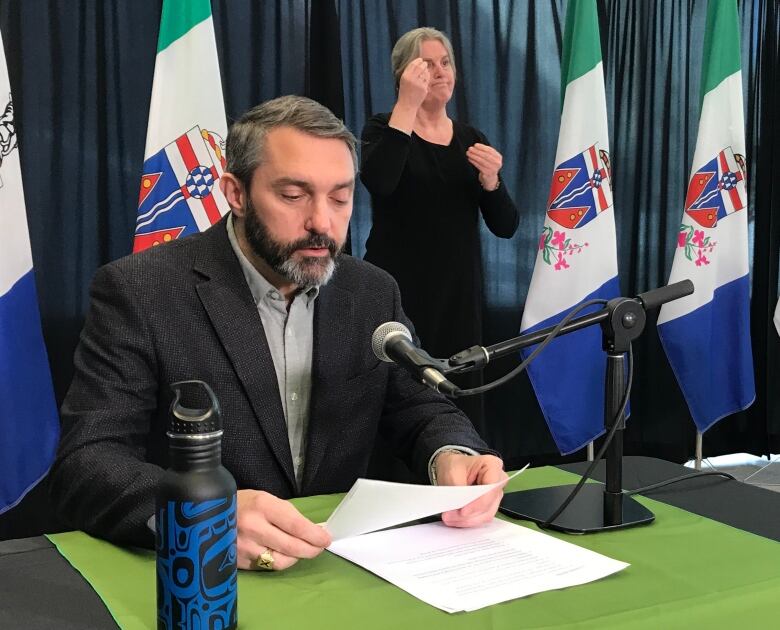 A man at a table sits before a microphone reading from some papers.