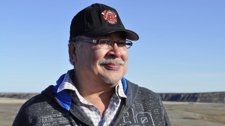 A man wearing a sweater a collared shirt and a cap looks off into the distance 