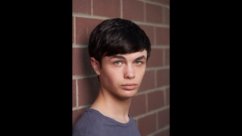 A young man with black hair and blue eyes, dressed in a grey-blue t-shirt is pictured in front of a brick wall, gazing intently at the camera.