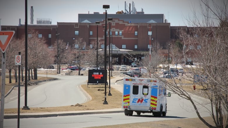 An ambulance drives down a curved road towards a red brick hospital building in the distance.