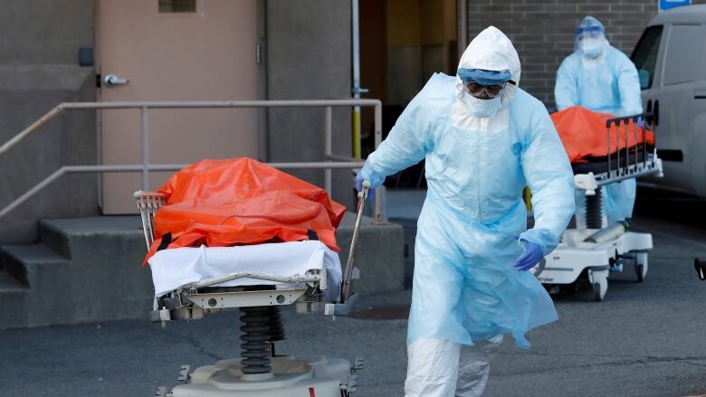 A person in heavy protective medical gear pulls a gurney bearing a body wrapped in orange plastic. 