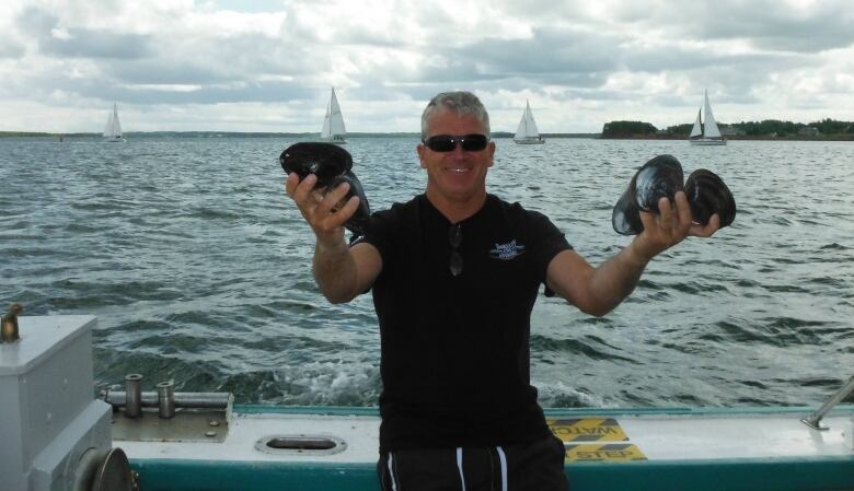 Perry Gotell sit on a boat in the water smiling holding two clams