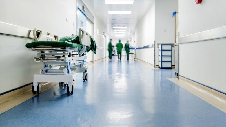 An empty gurney in the hallway of a hospital. Doctors in green scrubs stand in the background. 