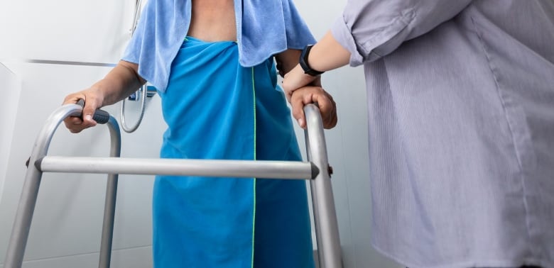 The hands of a person are on a walker. The left hand of another person is on that person's arm, supporting them.