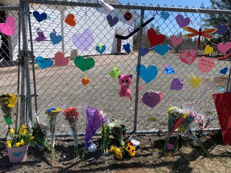 A chain link fence has colourful paper hearts and stuffed animals attached, with flowers and notes on the ground