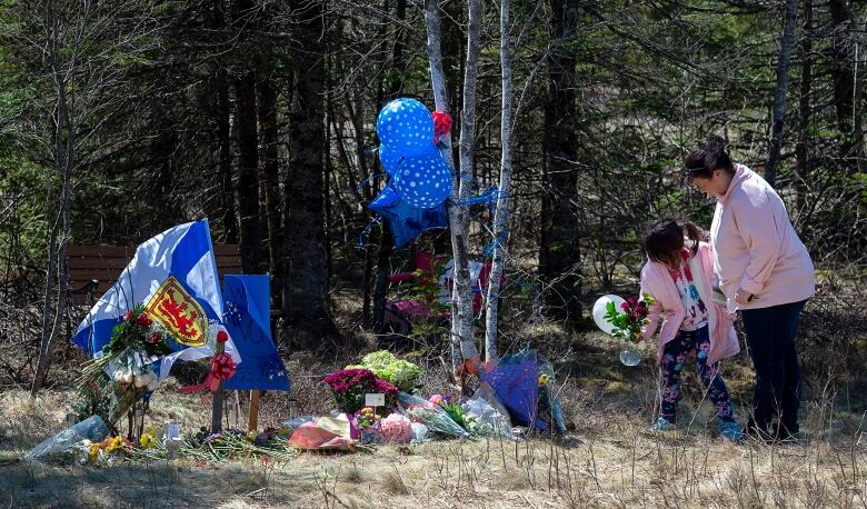 A woman and child stand near a grassy area where flags, balloons and flowers have been placed.