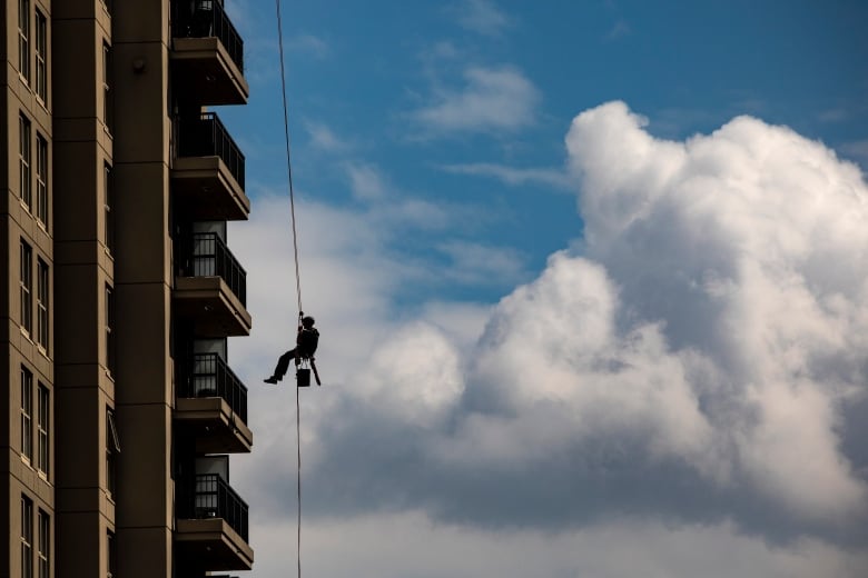 A man in a chair hangs from a skyscraper on a long rope with a bucket and squeegee in his hand.