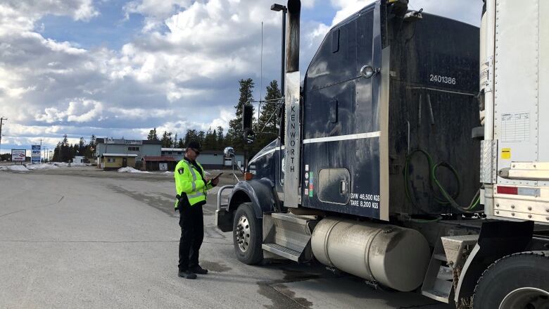 A person in a bright yellow safety jacket stands beside a transport truck at a highway checkstop.