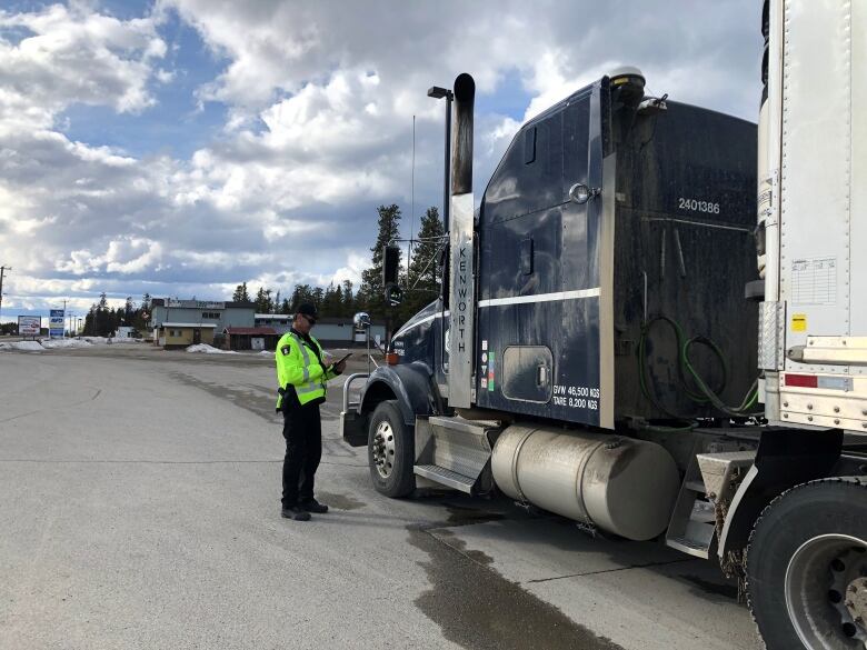 A person in a bright yellow safety jacket stands beside a transport truck at a highway checkstop.