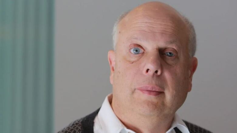 A man wearing a white button-up shirt with a black patterned cardigan sweater looks directly at the camera.