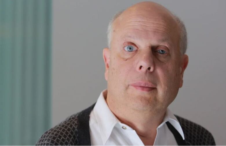 A man wearing a white button-up shirt with a black patterned cardigan sweater looks directly at the camera.