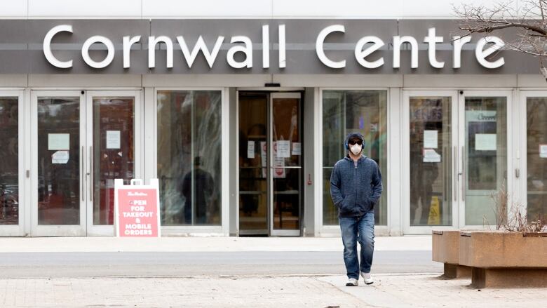 A man in blue jeans and a hoodie and wearing a mask walks away from the Cornwall Centre in Regina, Sask. 