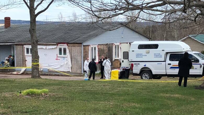 A home without siding surrounded by yellow police tape and police officers, some in hazmat suits. 