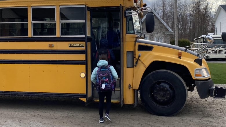 A child in a blue coat and purple backpack boards a yellow school bus 