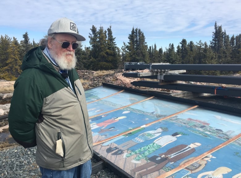 Bearded man stands beside mural lying faceup on gravel