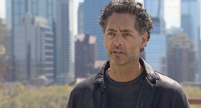 A man wearing a dark leather jacket stands with Calgary's skyline behind him.